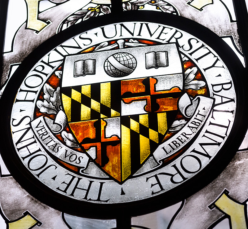 Image of a stained-glass window section on the Homewood campus showing the JHU shield and the Latin words Veritas Vos Liberabit.
